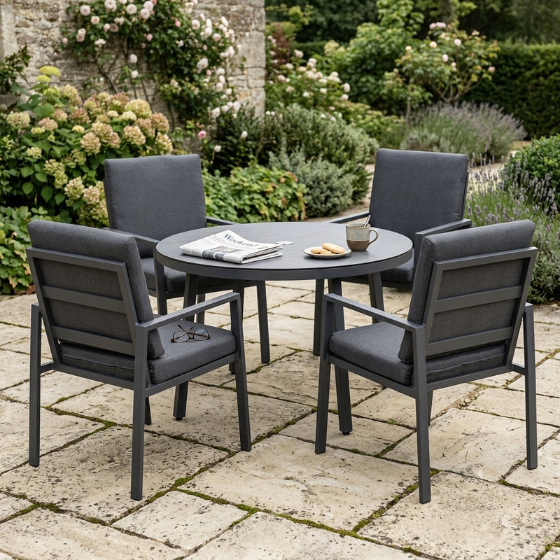 A 5-piece outdoor patio dining set featuring a round dark gray table and four matching armchairs with dark gray cushions, arranged on a stone patio in a garden setting.