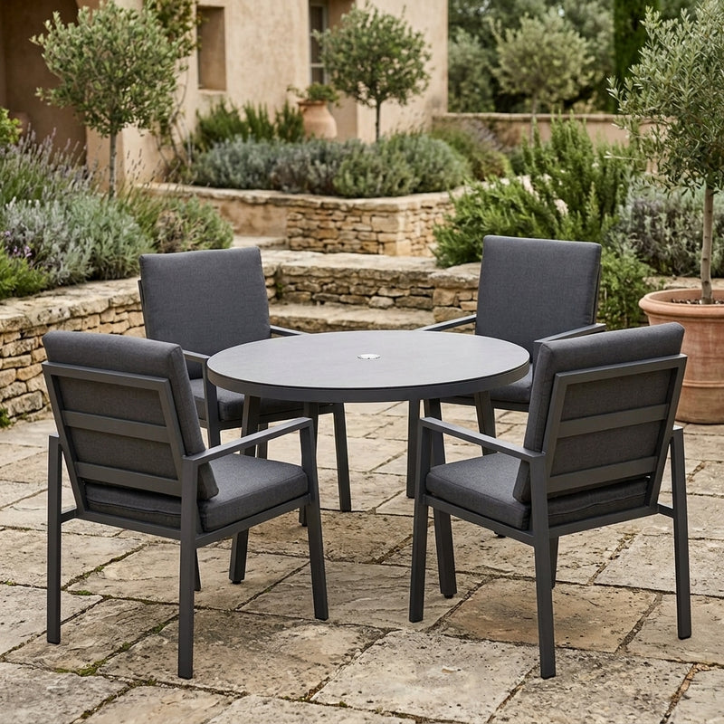 A 5-piece outdoor patio dining set featuring a round dark gray table and four matching armchairs with dark gray cushions, set on a stone patio in a garden environment.