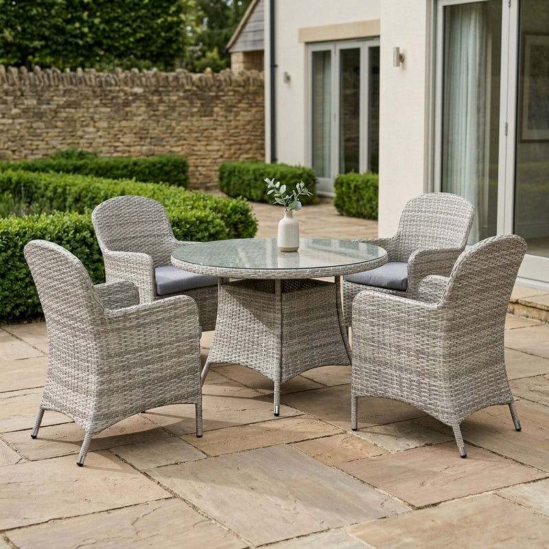 A 5-piece outdoor patio dining set featuring a round glass-top table and four woven wicker armchairs with gray cushions, set on a stone patio in front of a house.