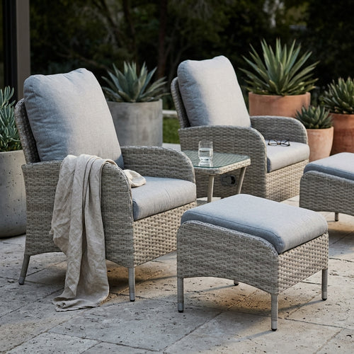 Gray wicker outdoor patio furniture set with light gray cushions, including two armchairs, a matching ottoman, and a small glass-top side table, arranged on a stone patio with potted plants in the background.