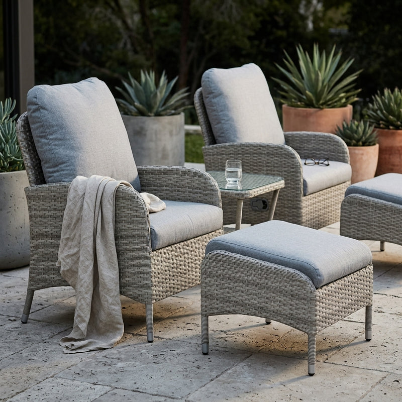 Gray wicker outdoor patio furniture set with light gray cushions, including two armchairs, a matching ottoman, and a small glass-top side table, arranged on a stone patio with potted plants in the background.
