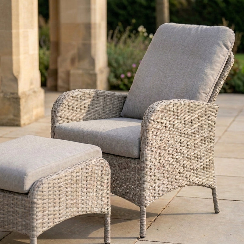 A light beige woven outdoor patio armchair with a matching ottoman and high back cushion, set on a stone patio in front of stone columns.