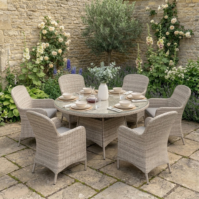A round outdoor dining set with a woven wicker table and six matching armchairs, styled on a stone patio with a garden backdrop featuring climbing roses and plants.