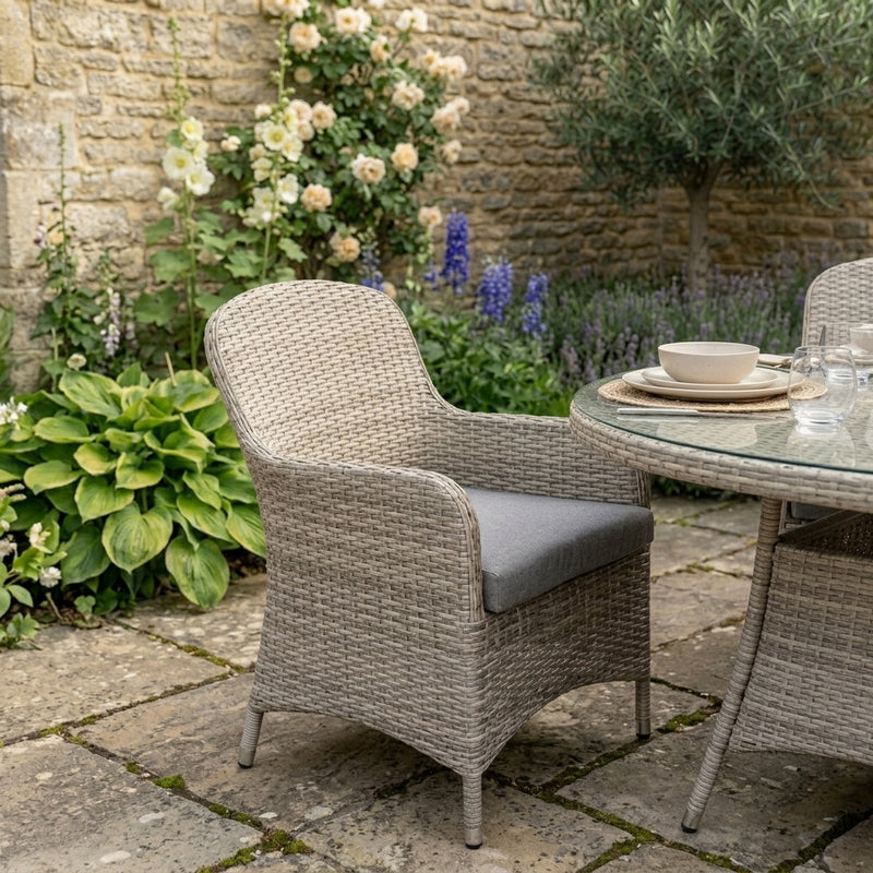 A woven outdoor patio armchair with a gray cushion sits beside a glass-top dining table on a stone patio, with flowers and a stone wall in the background.