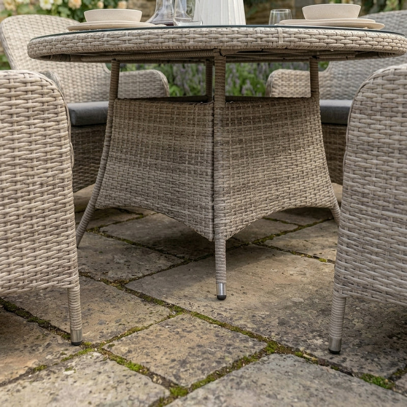 A round outdoor dining table set with woven wicker chairs on a stone patio. The table has a glass top and is surrounded by matching woven chairs with gray cushions.