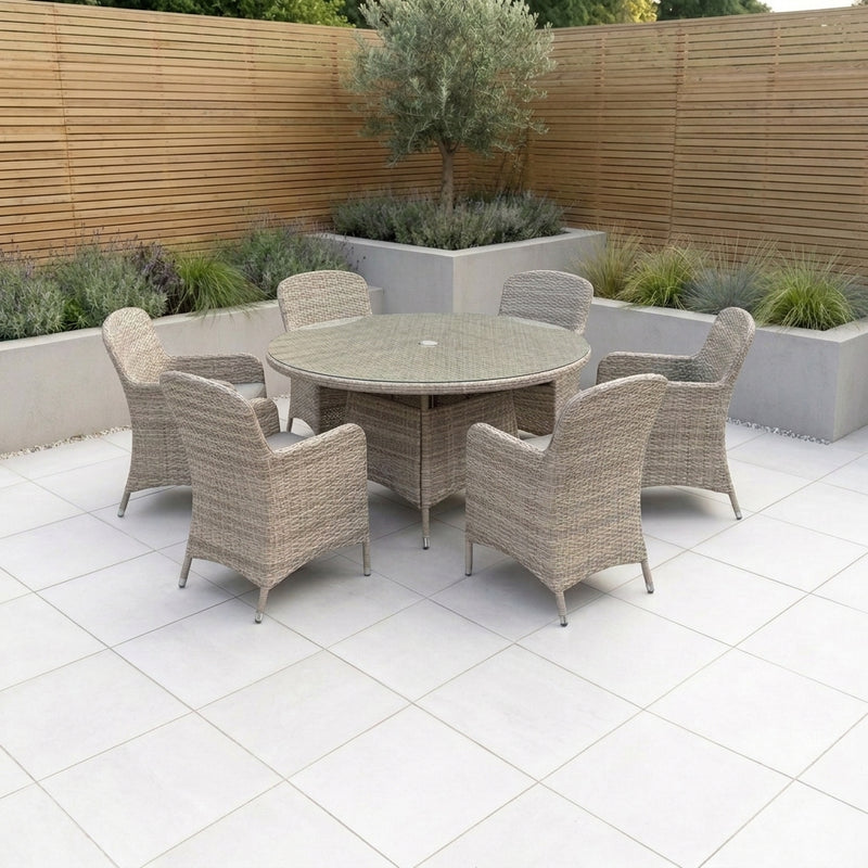 A 7-piece outdoor patio dining set featuring a round wicker table and six matching armchairs in a light beige wicker weave, set on a light tiled patio with a wooden slat fence and planter beds in the background.