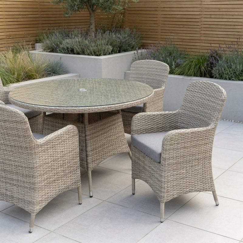 A 5-piece outdoor patio dining set featuring a round glass-top table and four woven wicker armchairs with light gray cushions, set on a tiled patio with garden plants in the background.