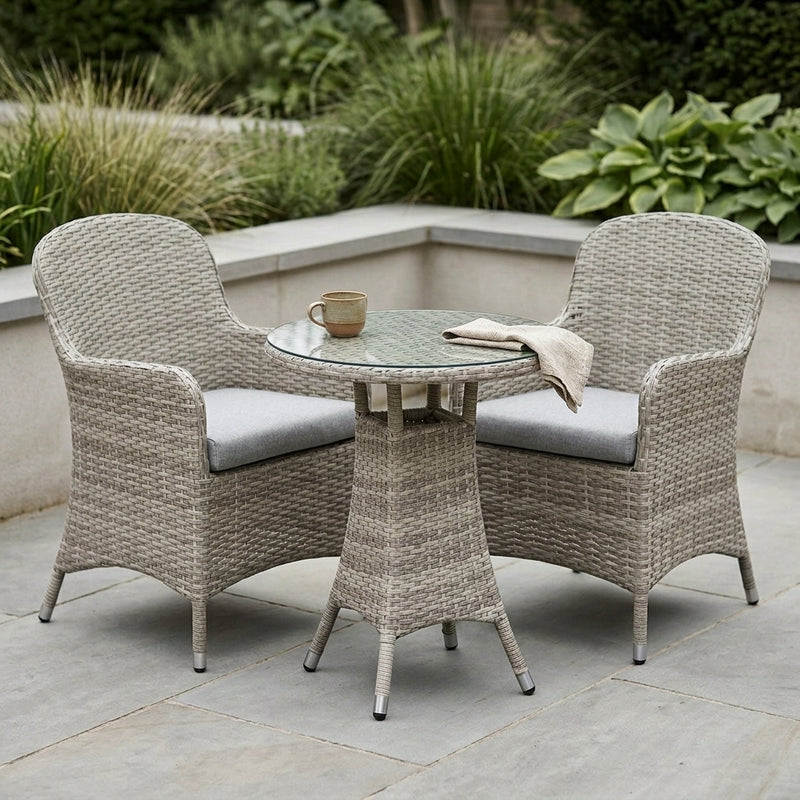 A 3-piece outdoor patio furniture set featuring two high-back armchairs with light gray cushions and a round glass-top side table, all in a woven taupe wicker material, set on a stone patio with green plants in the background.