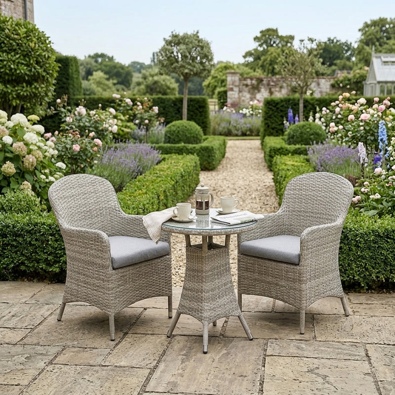 A 3-piece outdoor patio dining set featuring two woven wicker armchairs with light gray cushions and a matching round wicker coffee table, set on a stone patio in a beautifully landscaped garden.