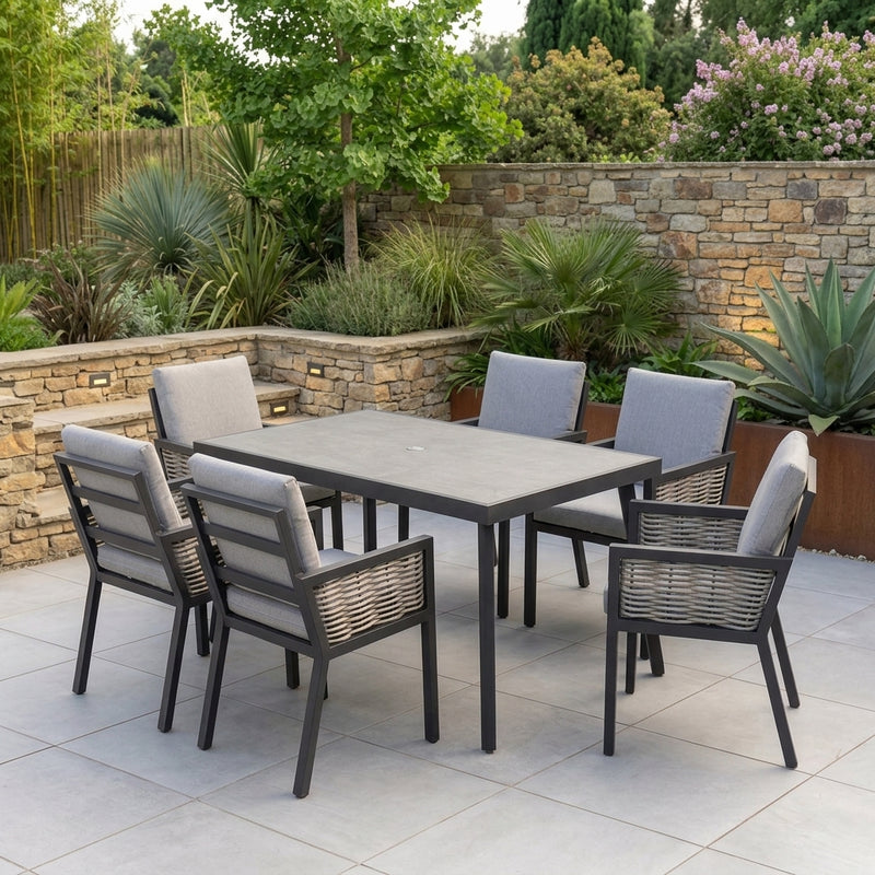A 7-piece outdoor patio dining set with a rectangular gray table and six gray cushioned chairs with dark gray frames, set on a light stone patio with a stone wall and green plants in the background.