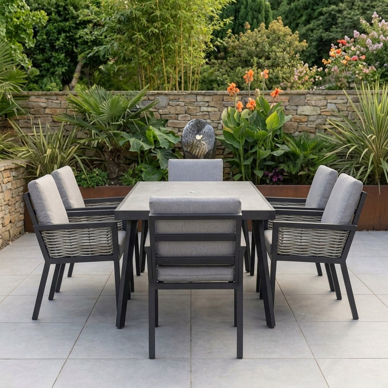 An outdoor patio dining set featuring a rectangular gray table and six matching armchairs with gray cushions, arranged on a light stone tile patio in front of a garden wall with plants.