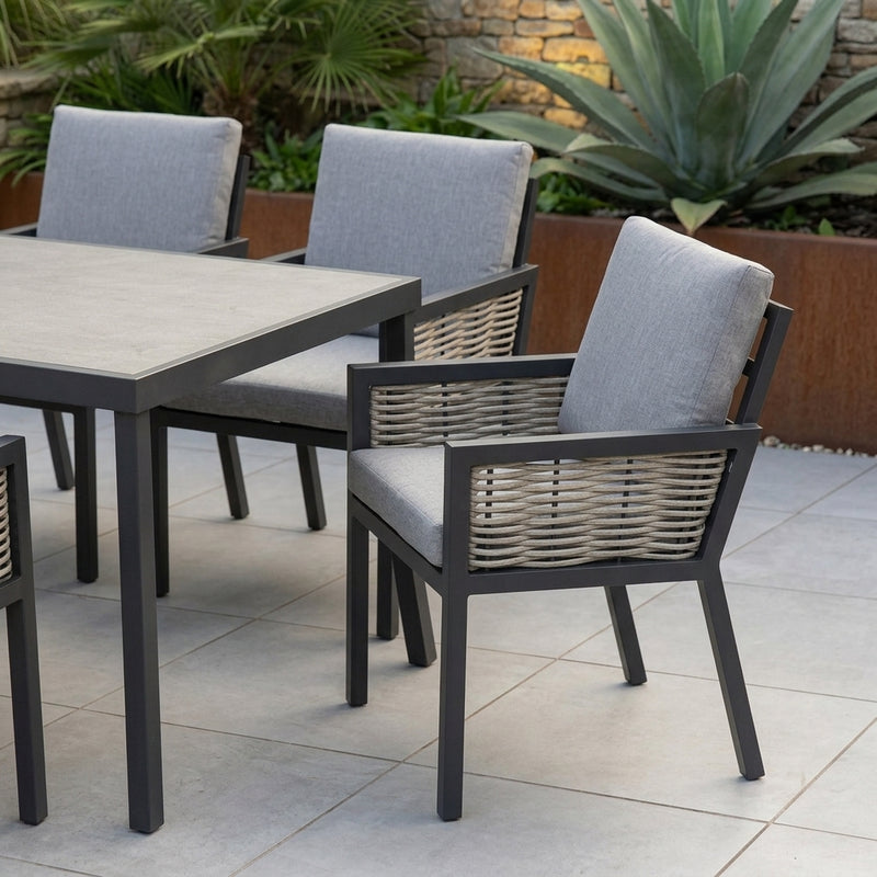 Outdoor patio dining set featuring a rectangular table with a light gray top and dark gray frame, surrounded by four armchairs with light gray cushions and woven wicker-style backs, set on a tiled patio with plants in the background.