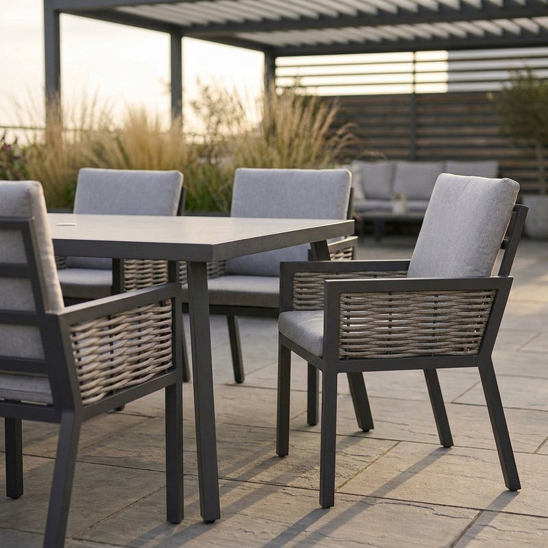 Outdoor dining set with a rectangular dark gray table and four matching armchairs with woven backs and light gray cushions, placed on a patio under a pergola.