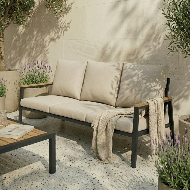 Beige outdoor sofa with cushions and a blanket on a patio with plants and a table.
